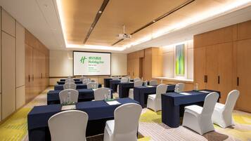 Sala de conferências com várias mesas, cadeiras brancas e uma tela de projeção exibindo uma apresentação.