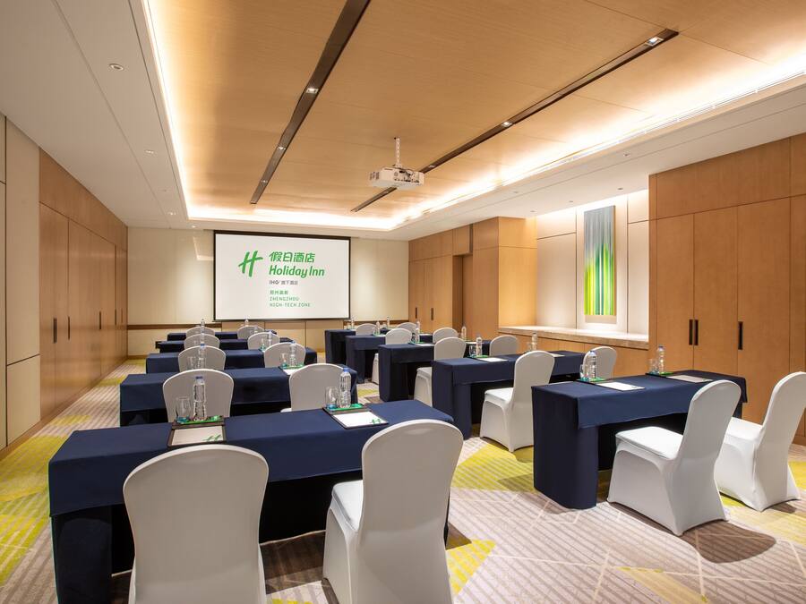 Sala de conferências com várias mesas, cadeiras brancas e uma tela de projeção exibindo uma apresentação.