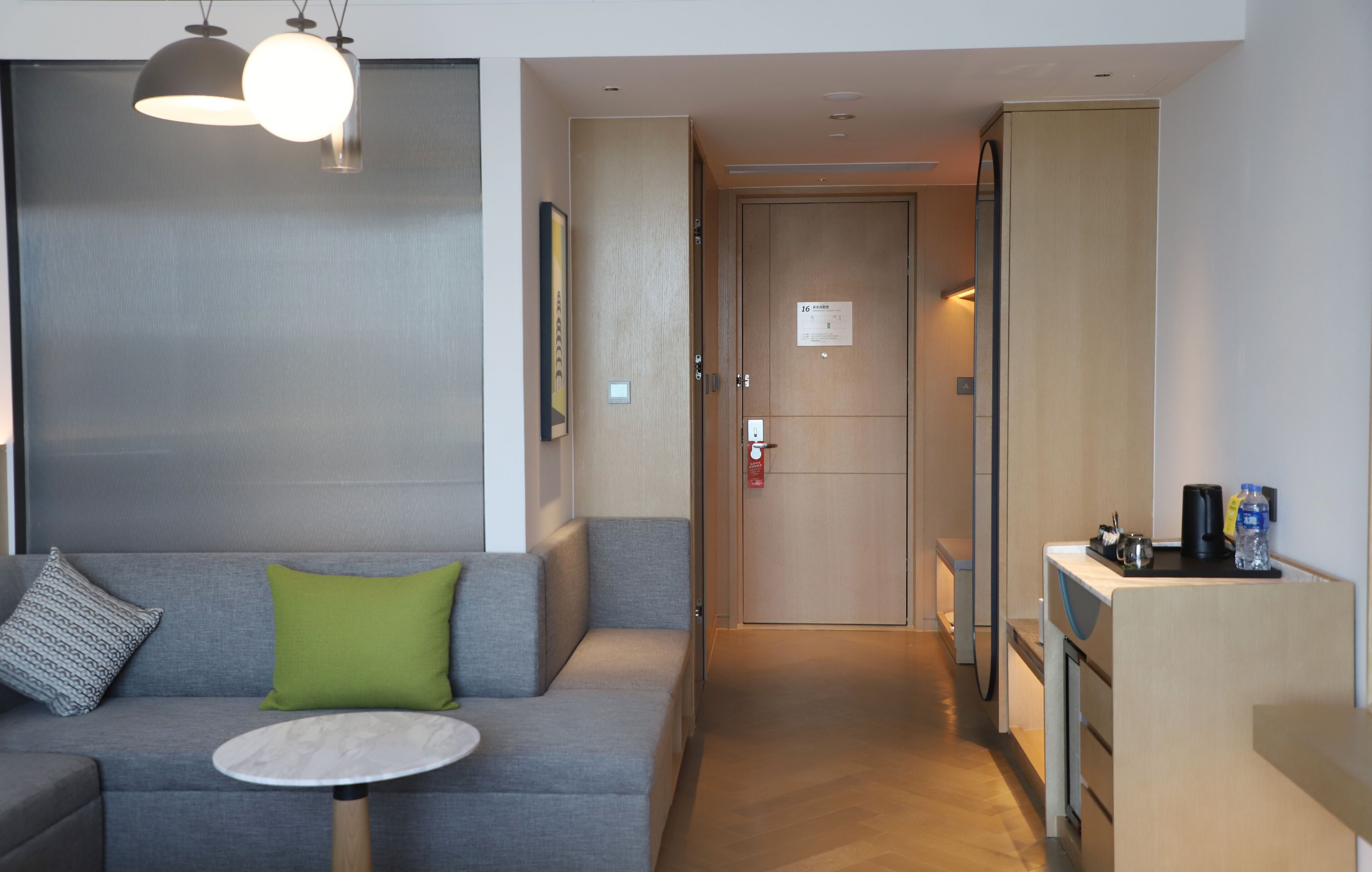 A modern hotel room with a grey sofa, a small round table, a green pillow, a wooden door, and a kitchenette area.