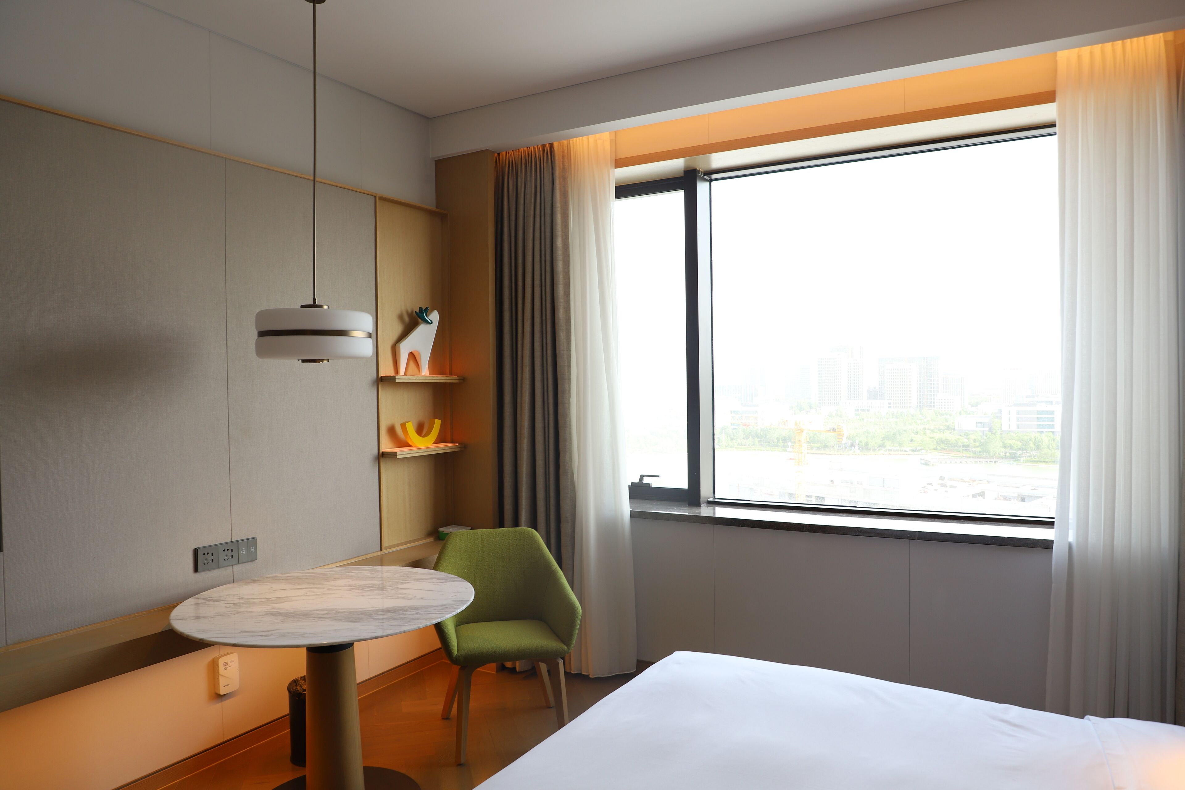 Une chambre d’hôtel avec un lit, une petite table ronde, une chaise verte, un luminaire suspendu et une fenêtre avec des rideaux.
