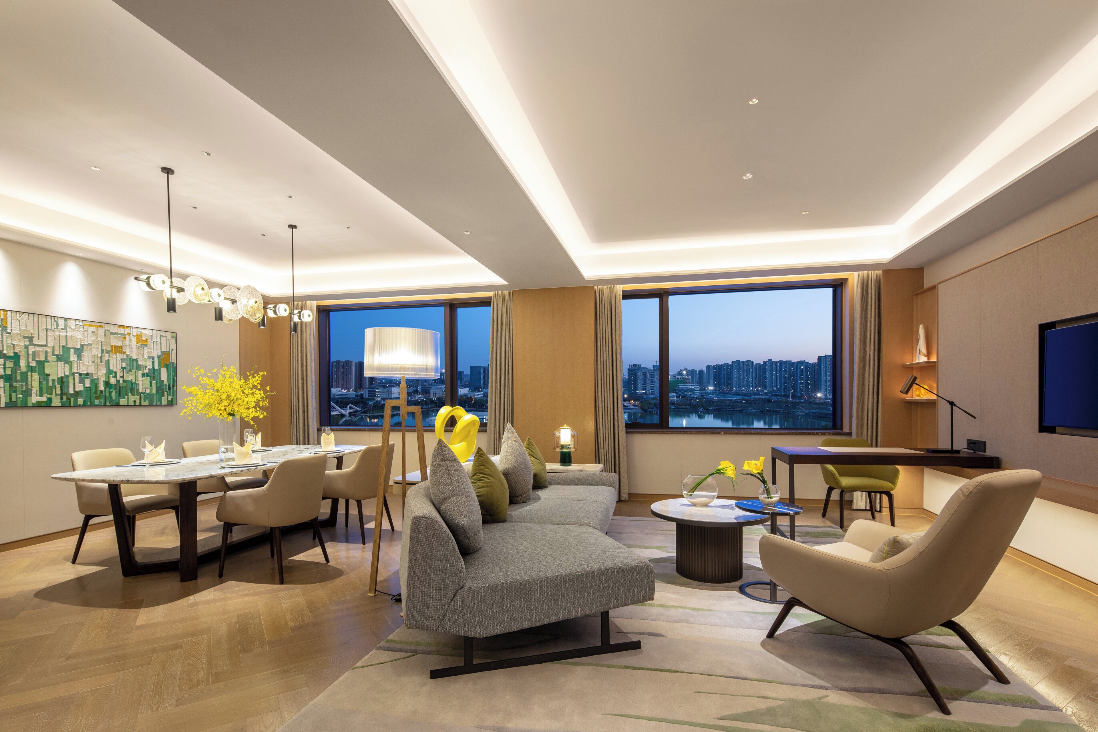 Une chambre d’hôtel moderne dotée d’un coin repas, d’un ensemble de canapés, d’une table basse et d’une grande fenêtre offrant une vue sur la ville.