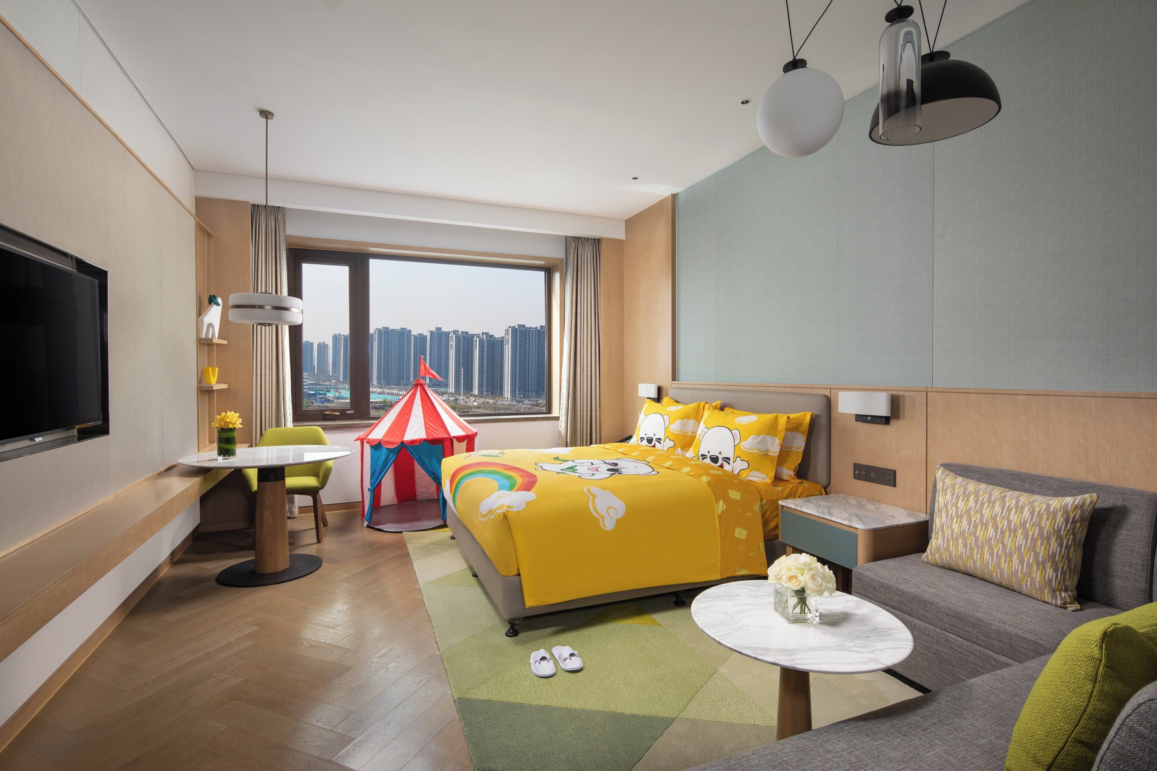 A hotel room with a bed, a desk, a TV, a window with a city view, and a small play tent.