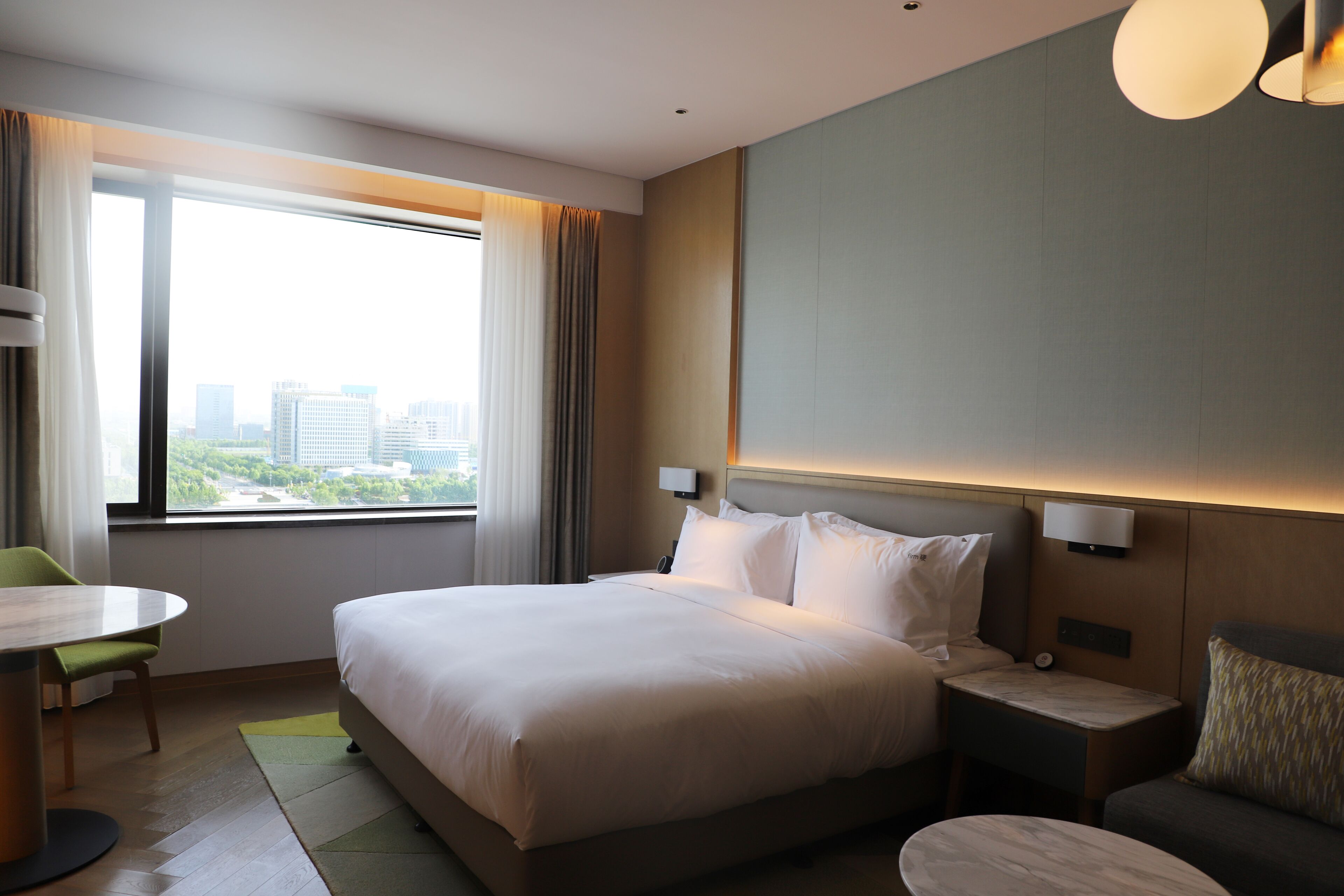 Ein Hotelzimmer mit einem großen Bett, einem Nachttisch, einem Sessel, einem kleinen Tisch und Blick auf die Stadt durch ein großes Fenster.