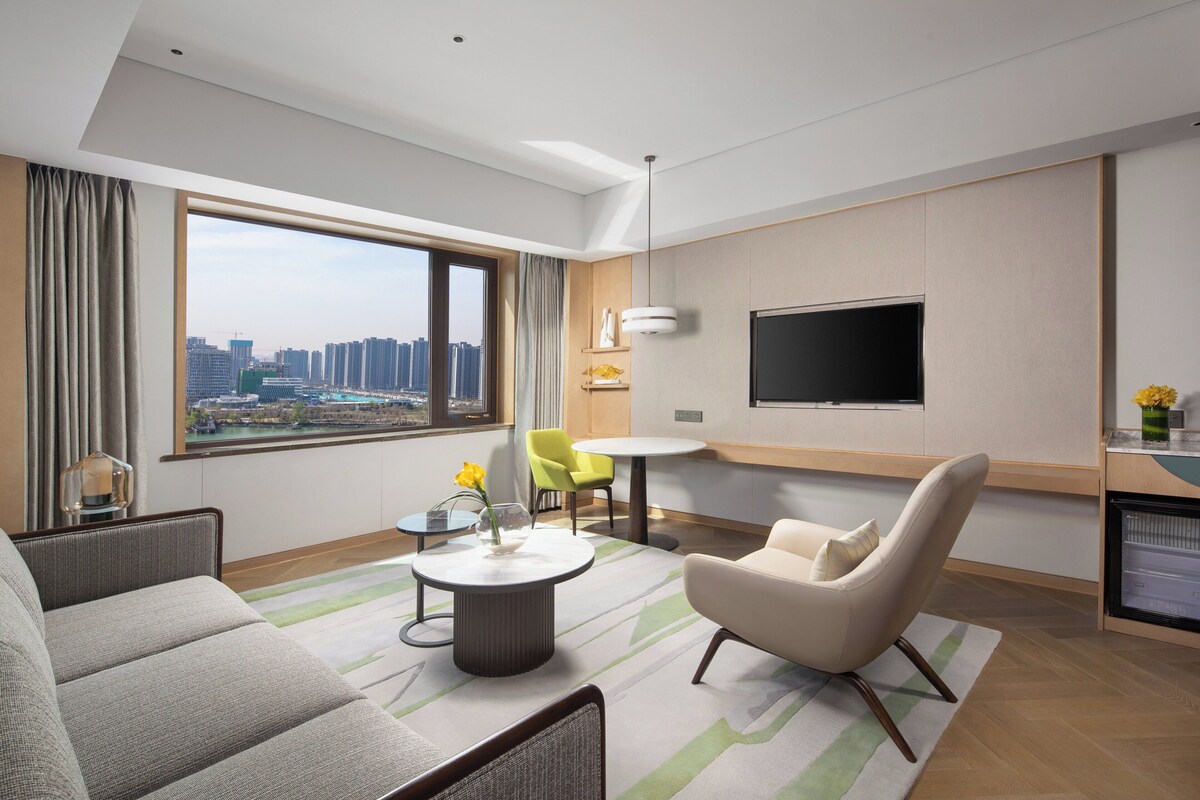 Ein modernes Hotelzimmer mit großem Fenster, Flachbildfernseher, Sofa, Sessel, kleinem Tisch und Minikühlschrank.