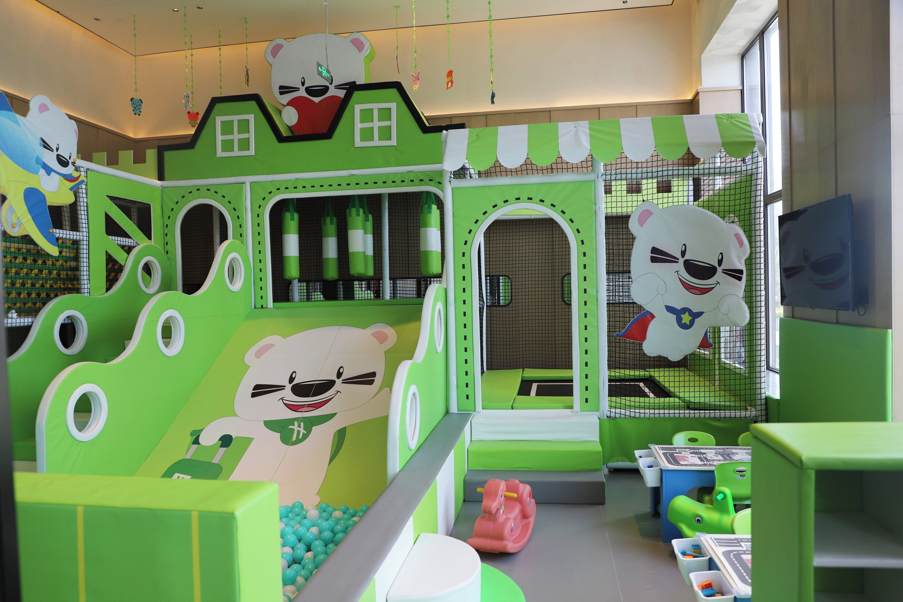 Un área de juegos infantiles con decoración verde y blanca, que incluye un tobogán con tema de oso y zonas de juego con redes.