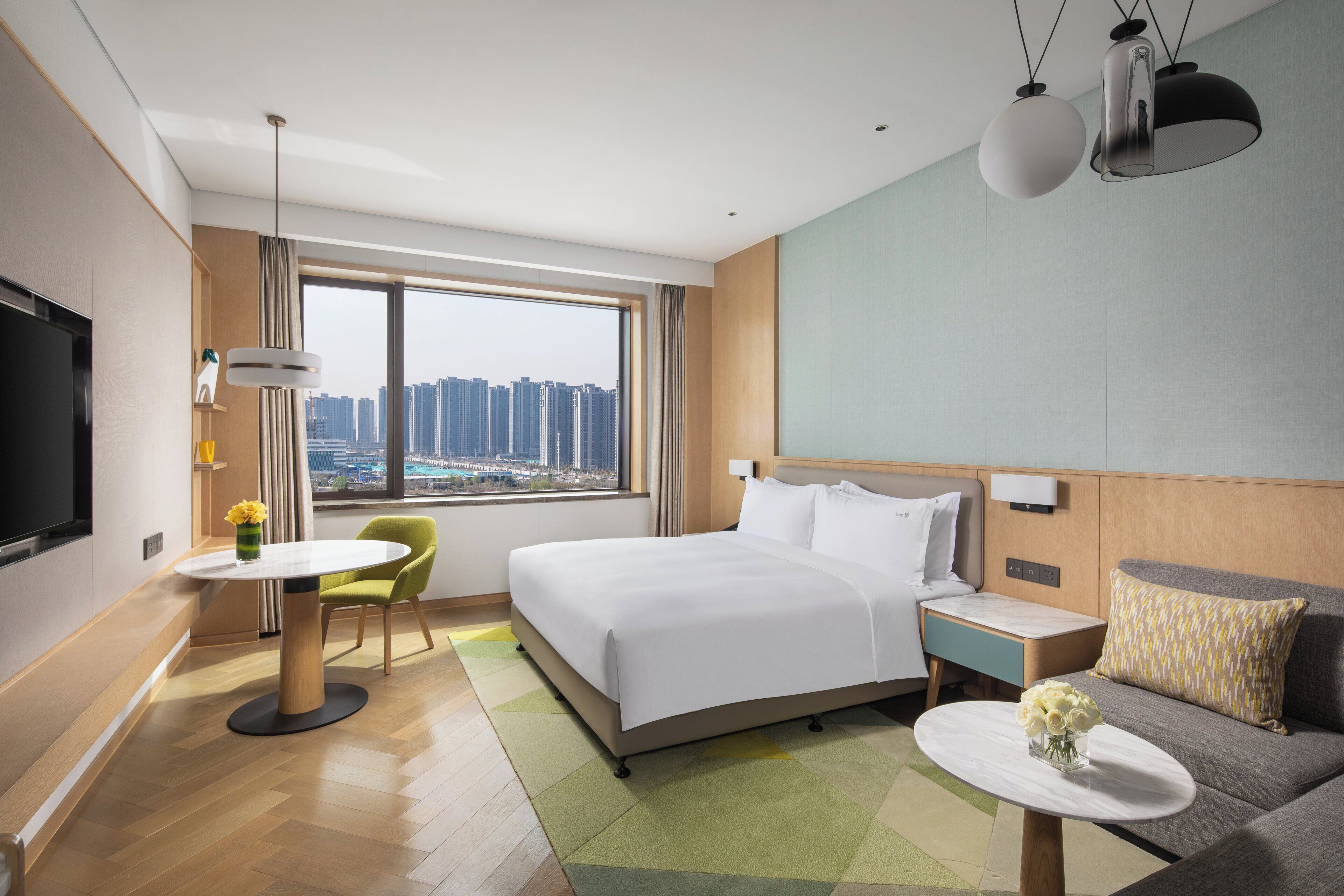 A hotel room with a bed, a sofa, a small table, a chair, a TV, and a window with a city view.