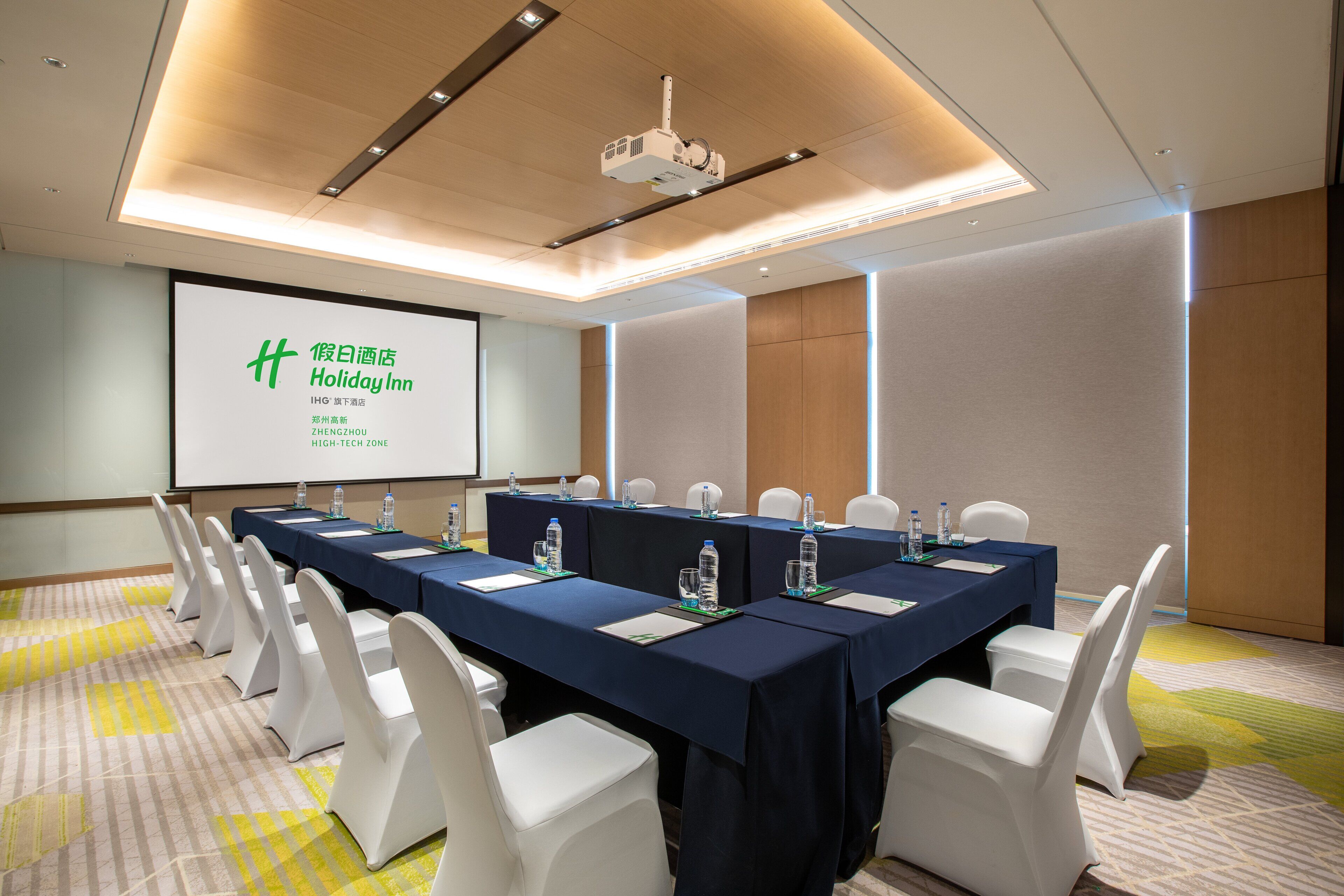 Uma sala de conferências com uma mesa comprida, cadeiras brancas e uma tela de projeção exibindo texto.