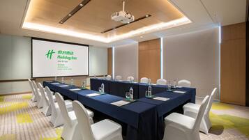 Uma sala de conferências com uma mesa comprida, cadeiras brancas e uma tela de projeção exibindo texto.