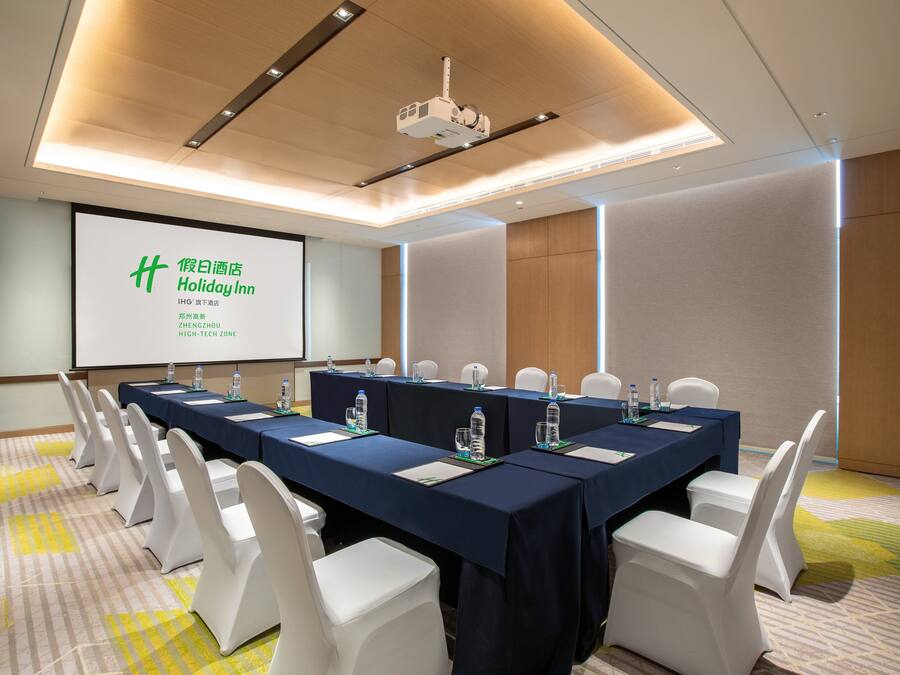 Uma sala de conferências com uma mesa comprida, cadeiras brancas e uma tela de projeção exibindo texto.