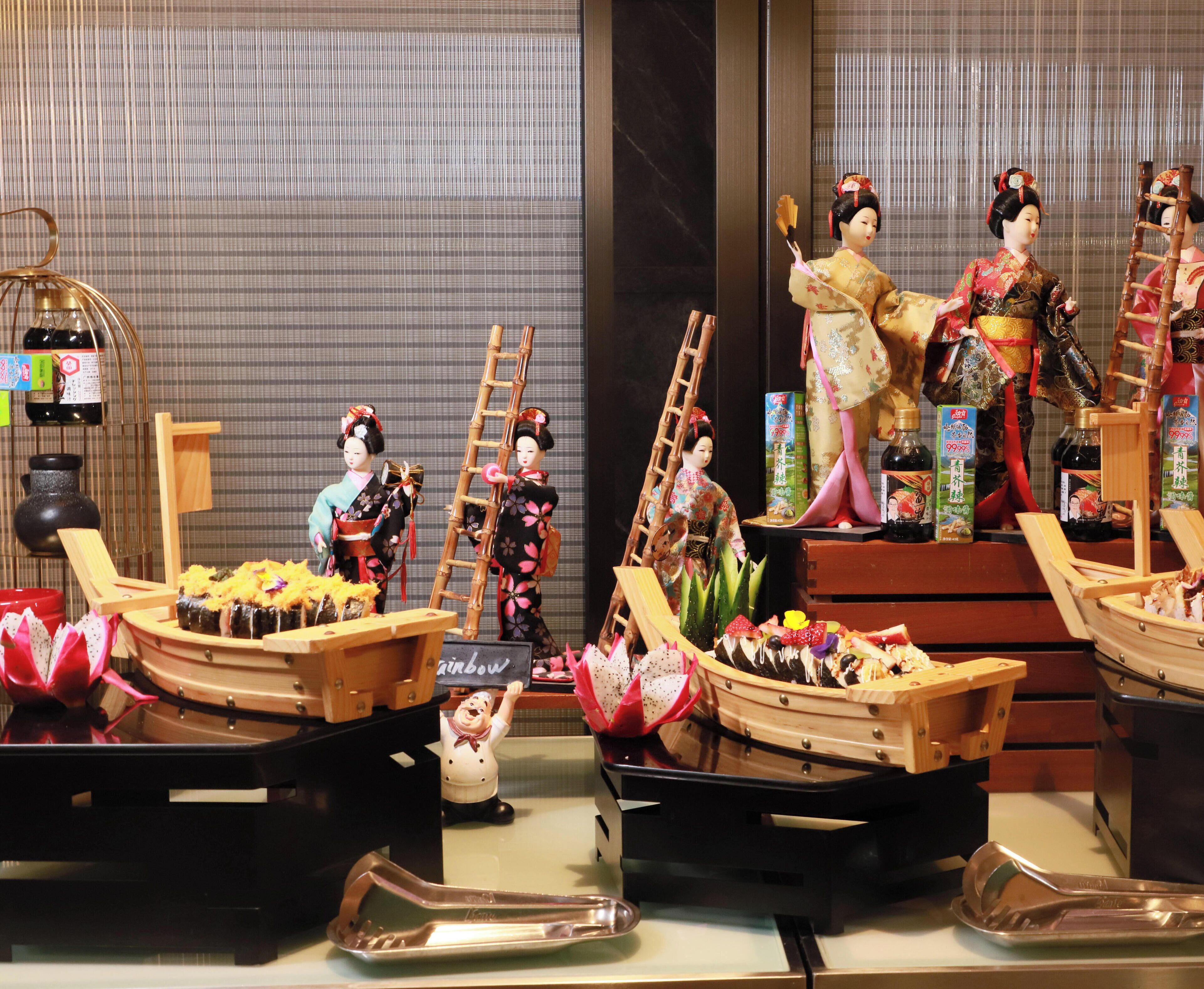 Una exhibición de muñecas japonesas tradicionales y alimentos, incluyendo sushi y botellas de sake, dispuestas sobre mesas.