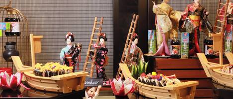 Una exhibición de muñecas japonesas tradicionales y alimentos, incluyendo sushi y botellas de sake, dispuestas sobre mesas.