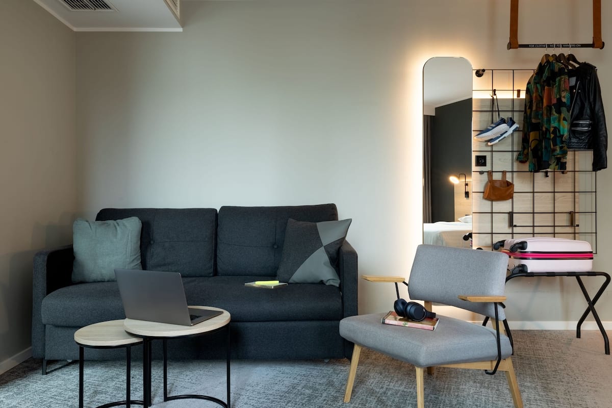 A modern living room with a grey sofa, a small round table with a laptop, a grey armchair with headphones, and a wall-mounted mirror with hanging clothes.