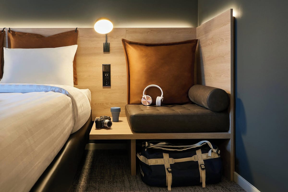A hotel room with a bed, a seating area with a pillow, a wall-mounted light, a small table with a camera and a cup, and a bag under the table.