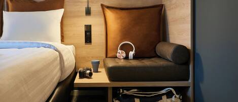 Une chambre d’hôtel avec un lit, un coin salon avec un coussin, une lampe fixée au mur, une petite table avec un appareil photo et une tasse, et un sac sous la table.