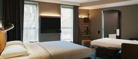 Habitación de hotel con una cama grande, una cama más pequeña, un escritorio, una silla, un televisor y una ventana con cortinas.