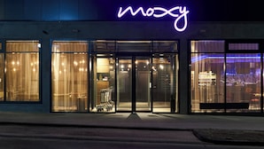 Un edificio con un letrero de neón que dice "moosy" y grandes puertas de vidrio que dan a un interior con muebles a la vista, y una banca en el exterior.