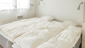 A bed with white bedding in a room with a window, a wall-mounted heater, and a wall-mounted light fixture.