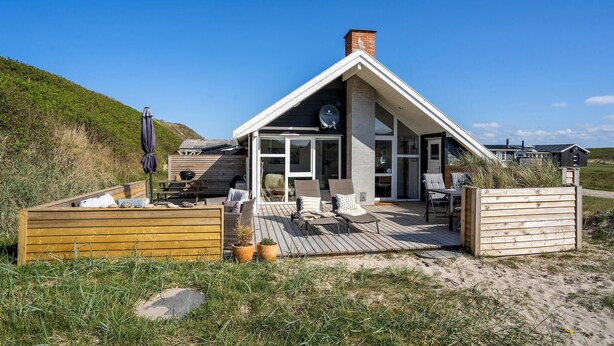 Une maison de plage dotée d’une terrasse en bois, de meubles de jardin et d’une grande fenêtre donnant sur la plage.