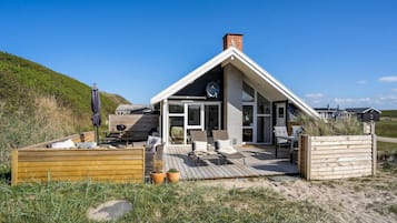 Et strandhus med træterrasse, udendørs møbler og et stort vindue med udsigt over stranden.