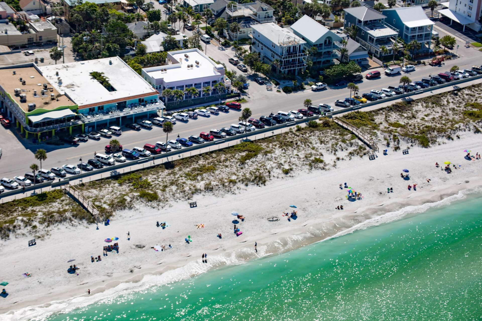 An aerial view of a coastal area with a beach, a road with parked cars, and buildings with balconies.