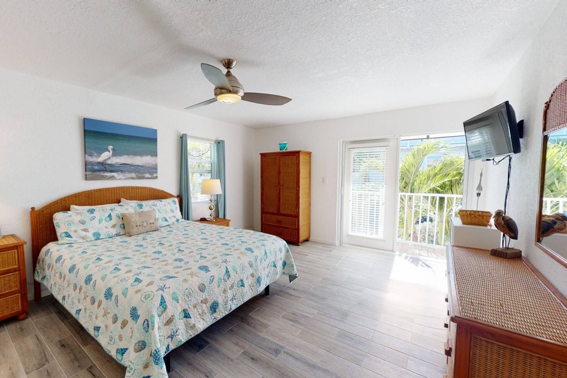 A bedroom with a bed, a ceiling fan, a painting, a wardrobe, a television, and a balcony with a view of greenery.