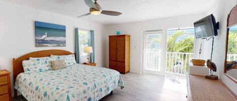 A bedroom with a bed, a ceiling fan, a painting, a wardrobe, a television, and a balcony with a view of greenery.
