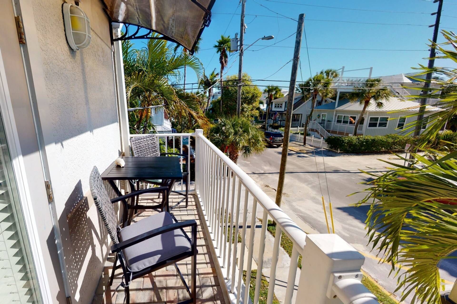 A balcony with a table and chairs, a view of a street with palm trees, and a building with a white facade.