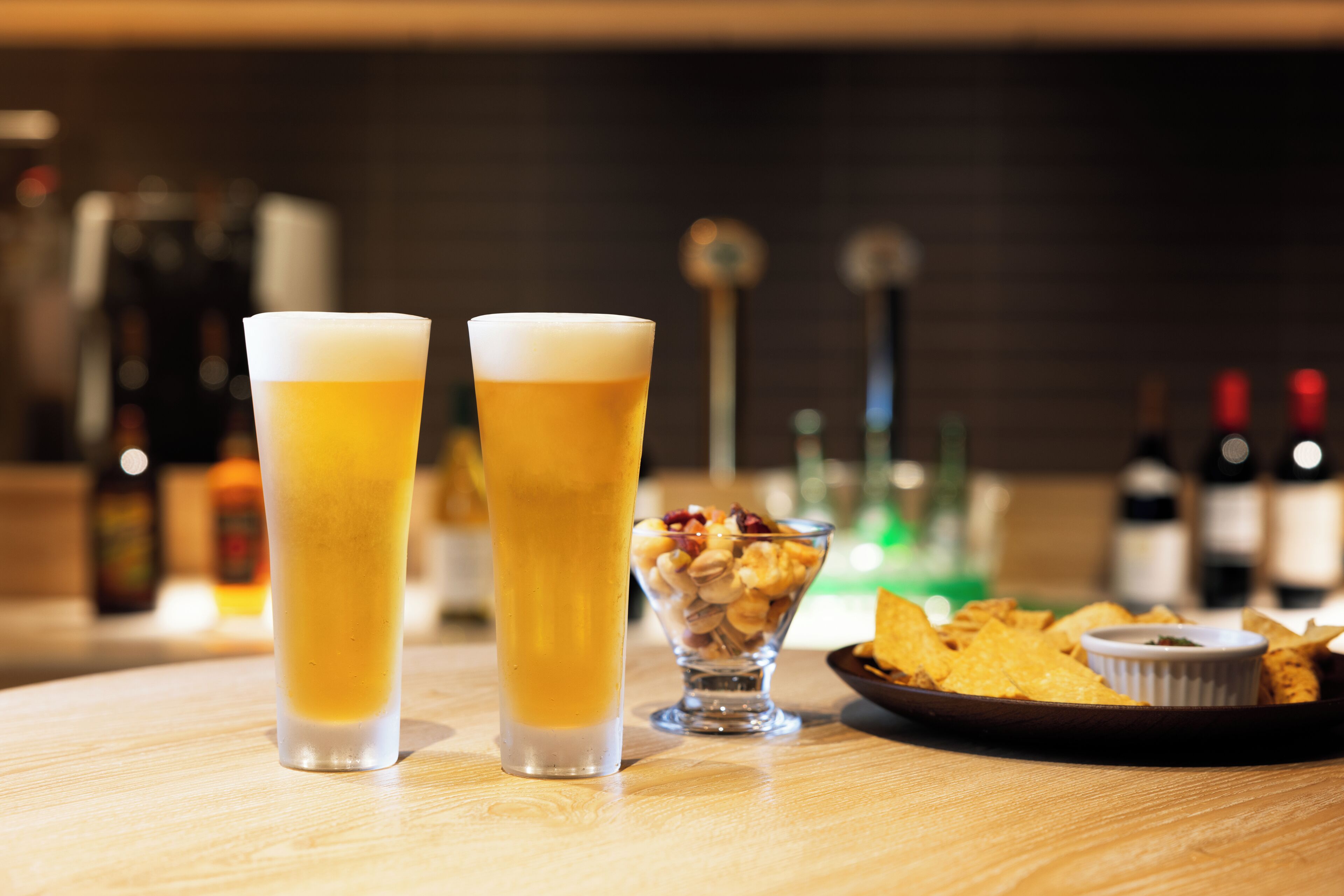 Deux grands verres de bière sur une table en bois, accompagnés d’un bol de noix et de chips, et une assiette de chips de tortillas avec une sauce pour tremper.