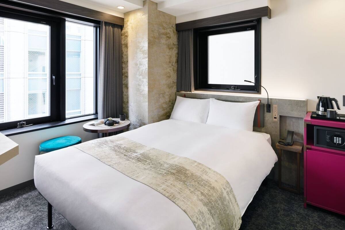 A hotel room with a large bed, a desk, a small round table, a minibar, and a window with curtains.