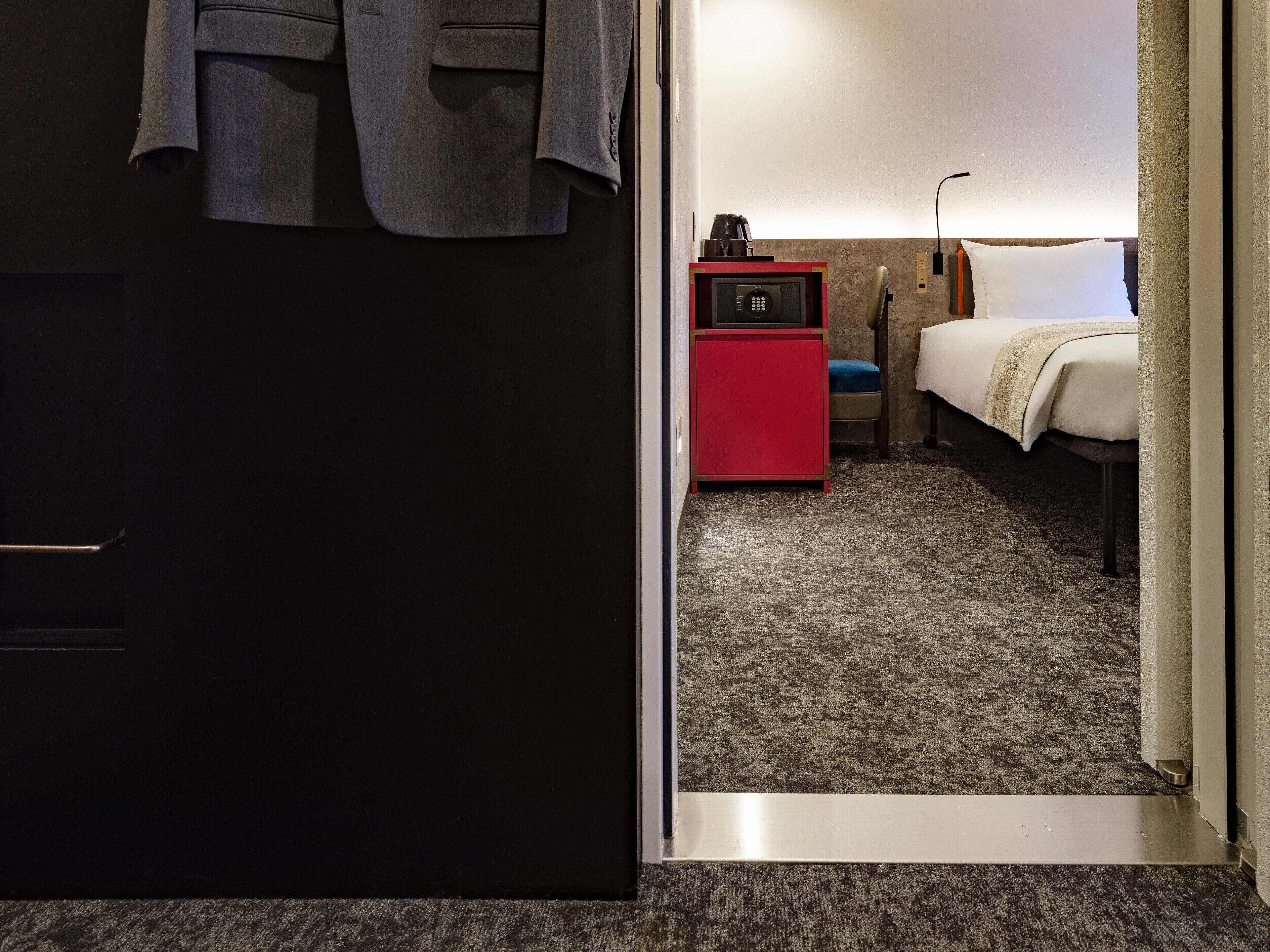 A hotel room with a bed, a red nightstand with a coffee maker, and a door leading to another room.