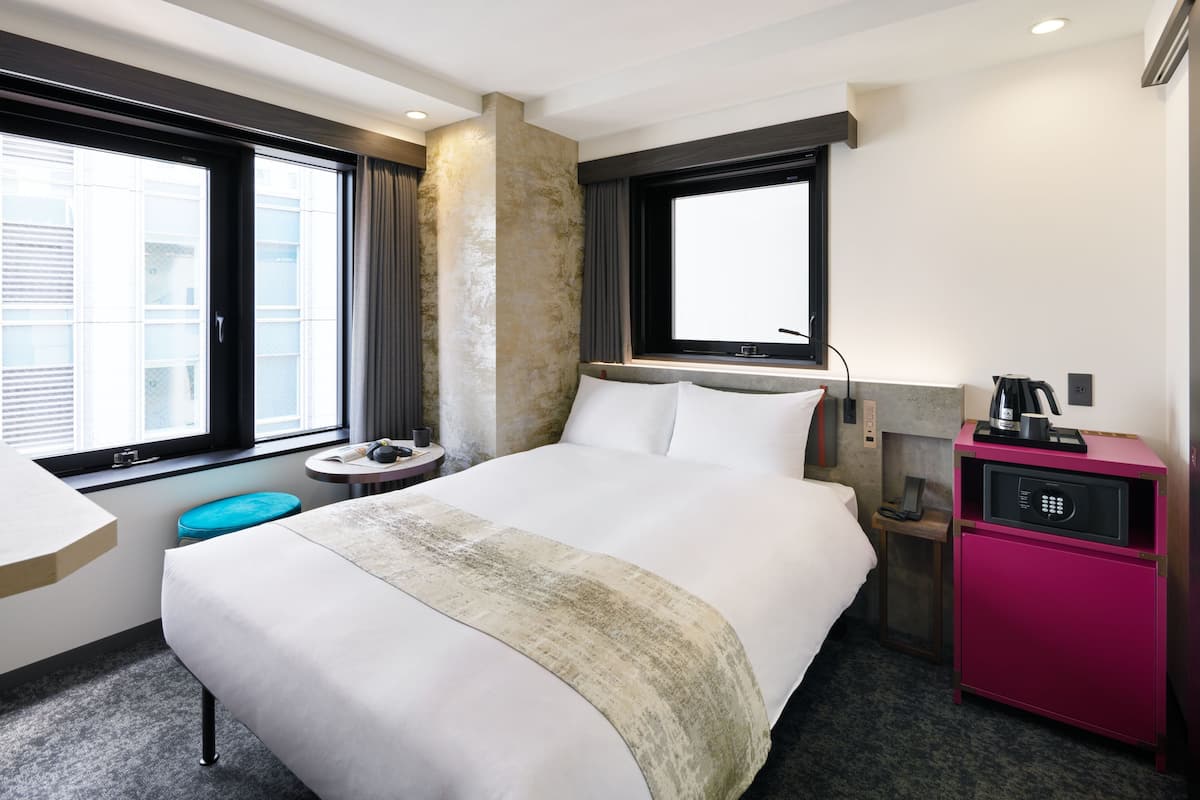 A modern hotel room with a large bed, a small desk, a minibar, and a view of the city through large windows.