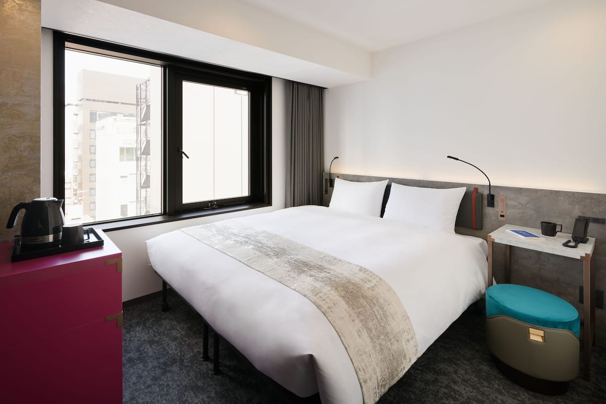 A hotel room with a large bed, a desk with a lamp, a coffee maker, and a view of the city through the window.