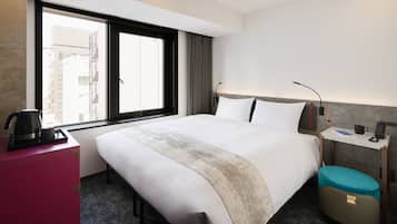 Habitación de hotel con una cama grande, un escritorio con lámpara, cafetera y vista a edificios a través del ventanal.