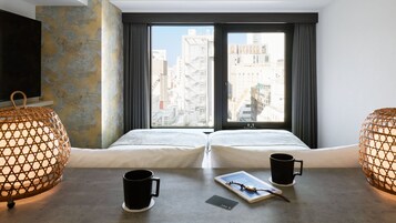 A hotel room with two beds, a large window, and a view of the city. There are two black mugs on the bedside table.