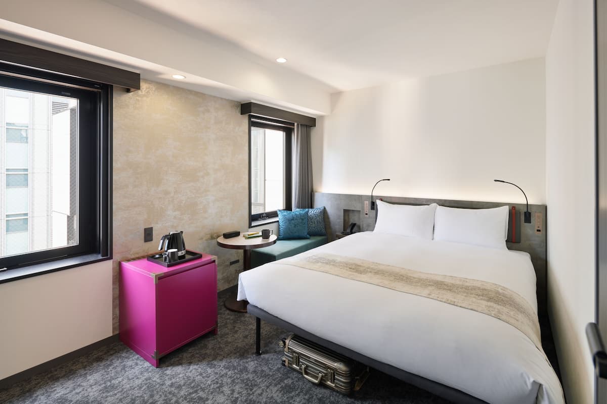 A hotel room with a bed, a small desk, a pink cabinet, a window, and a view of a building.
