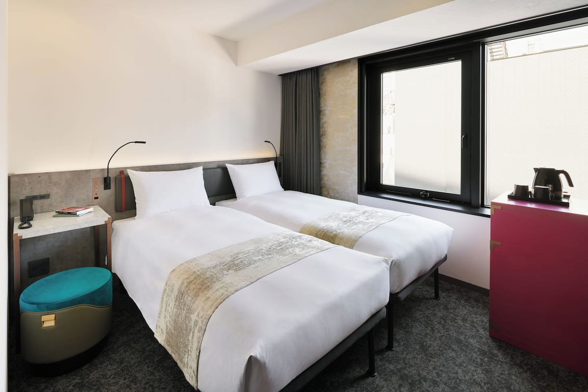 A hotel room with two beds, a desk with a coffee maker, a small ottoman, and a red cabinet.