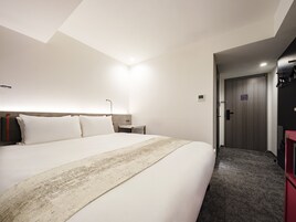 Hotellihuone, jossa on suuri sänky, yöpöytä, pieni sivupöytä, puinen ovi ja matolla peitetty lattia.