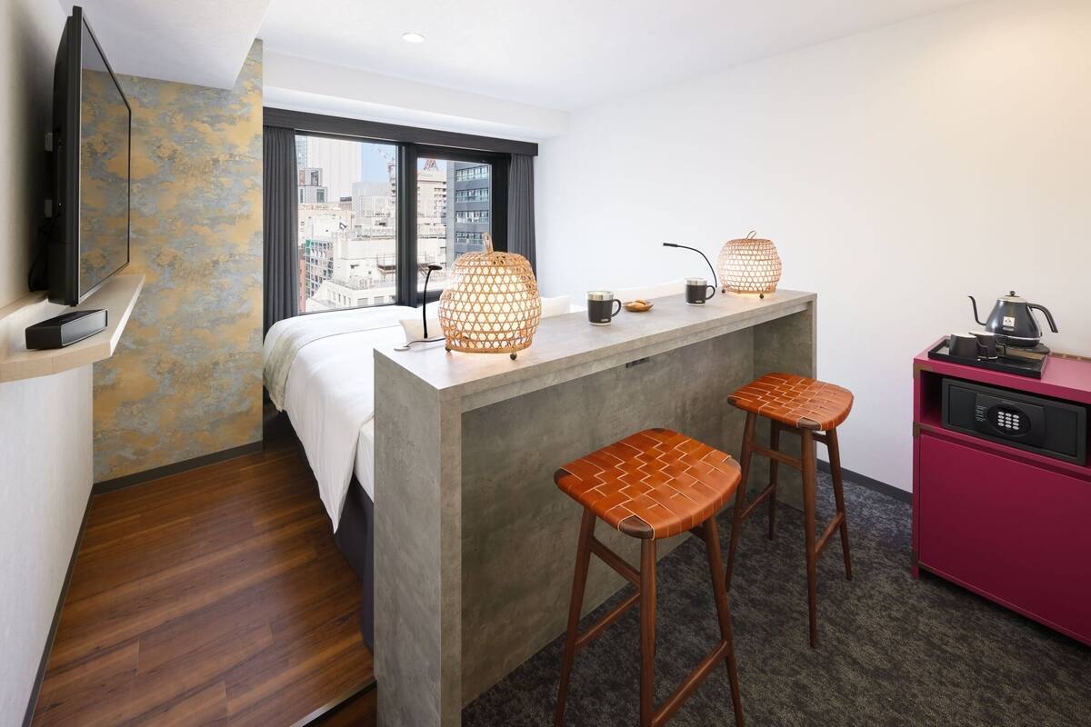 A compact hotel room with a bed, a small kitchenette, a TV, and a city view.