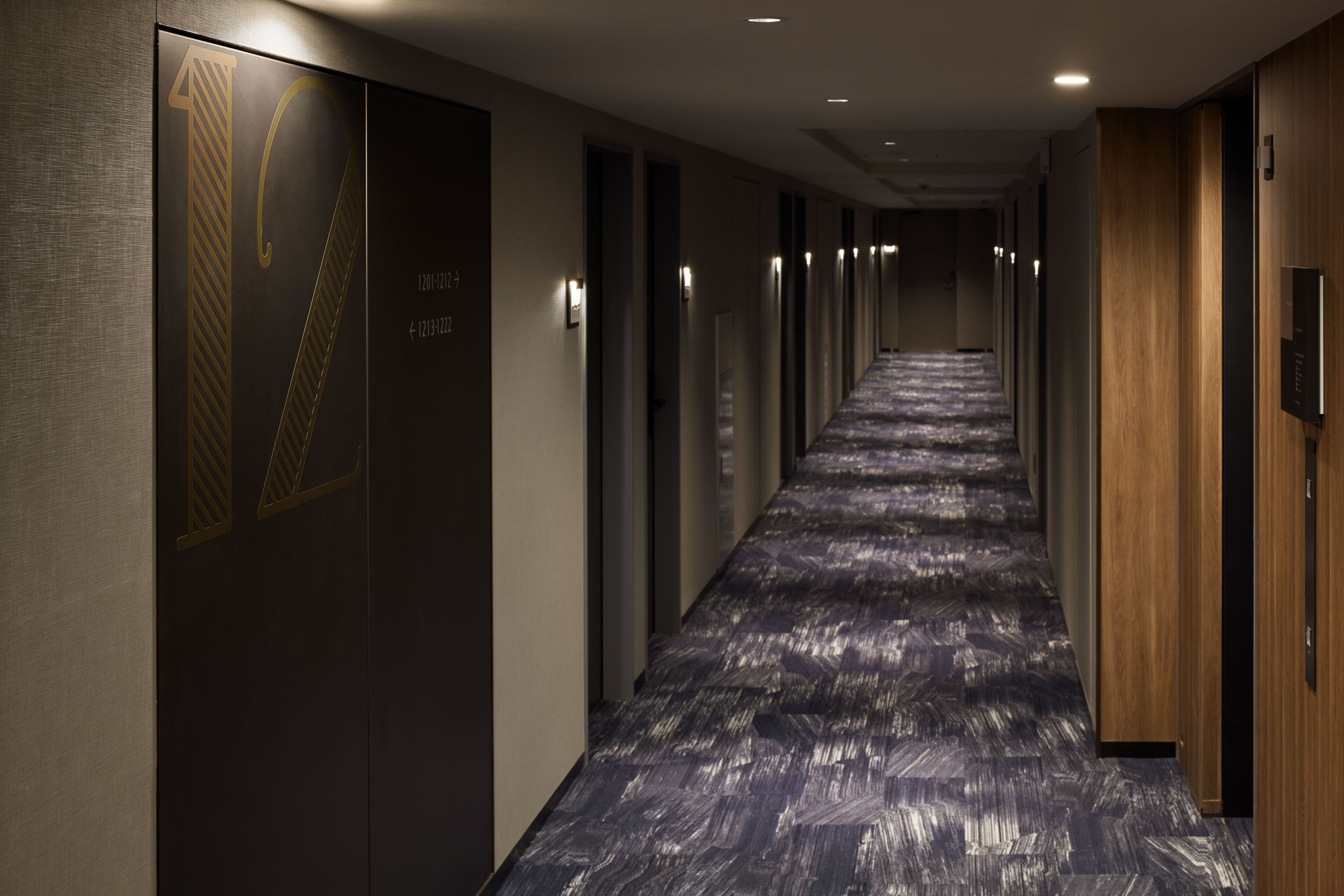 A long, narrow hotel corridor with a patterned carpet, wooden paneling on the right, and doors with numbers on the left.