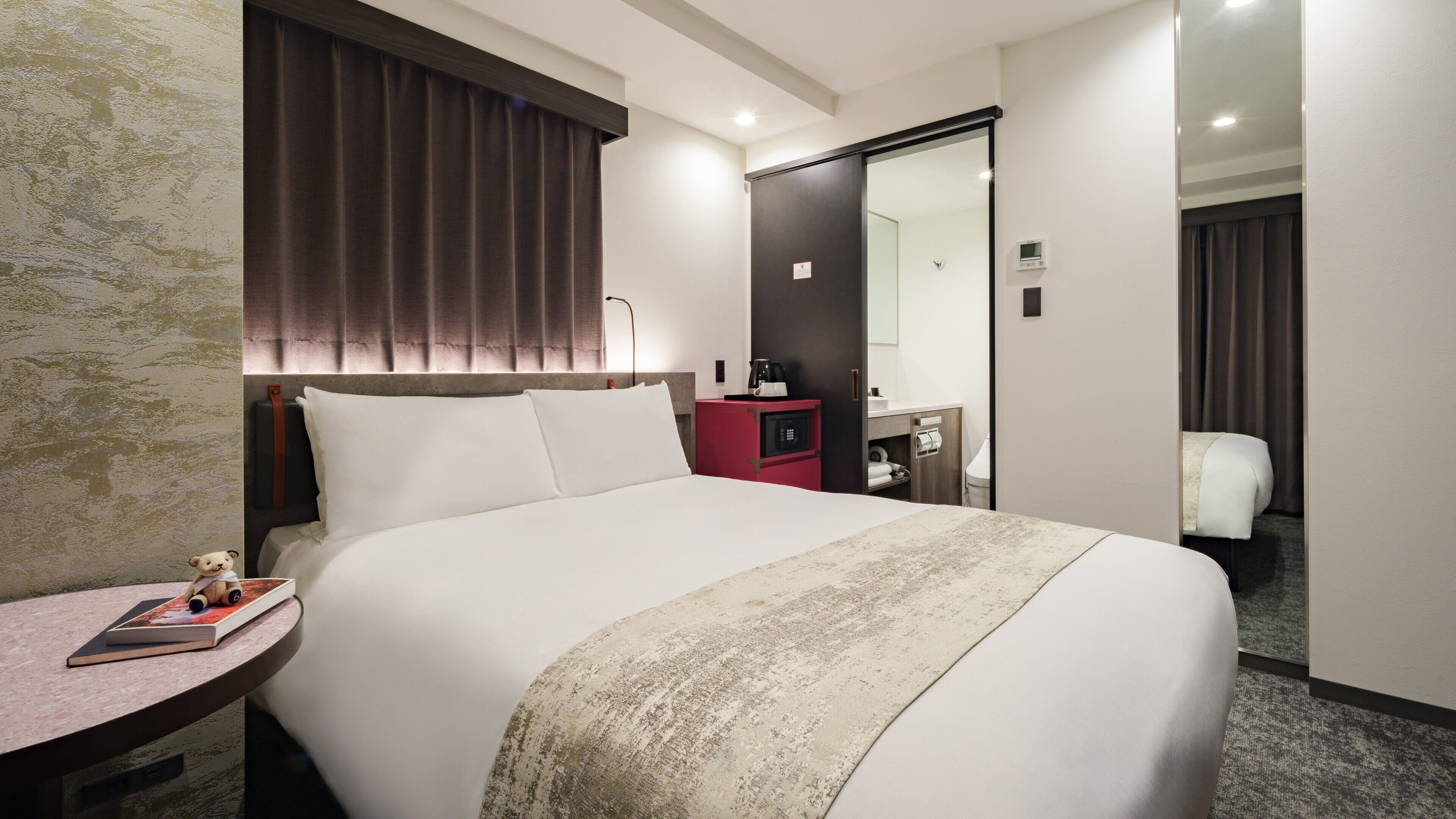 A hotel room with a large bed, a bedside table with a book and a teddy bear, a red minibar, and a bathroom visible through an open door.