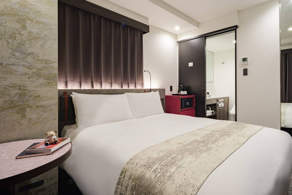 A hotel room with a large bed, a bedside table with a book and a teddy bear, a red minibar, and a bathroom visible through an open door.