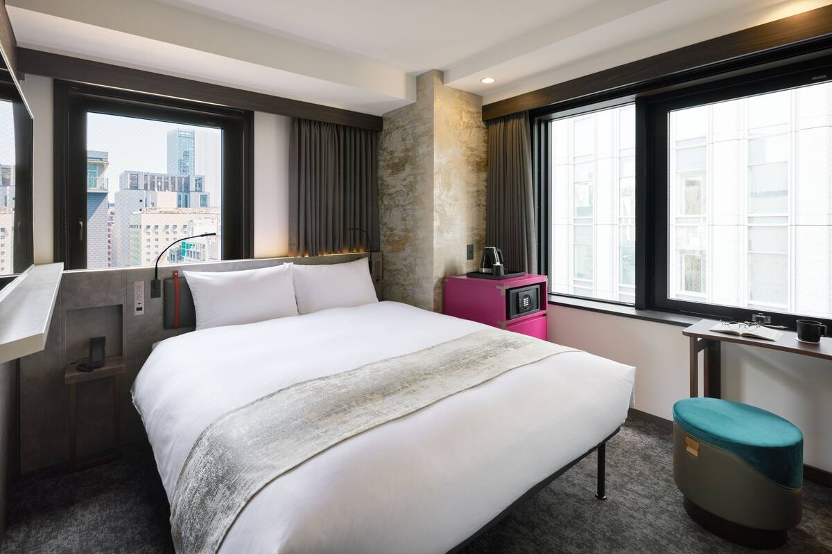 A hotel room with a large bed, a desk, a chair, a window with a city view, and a small seating area.