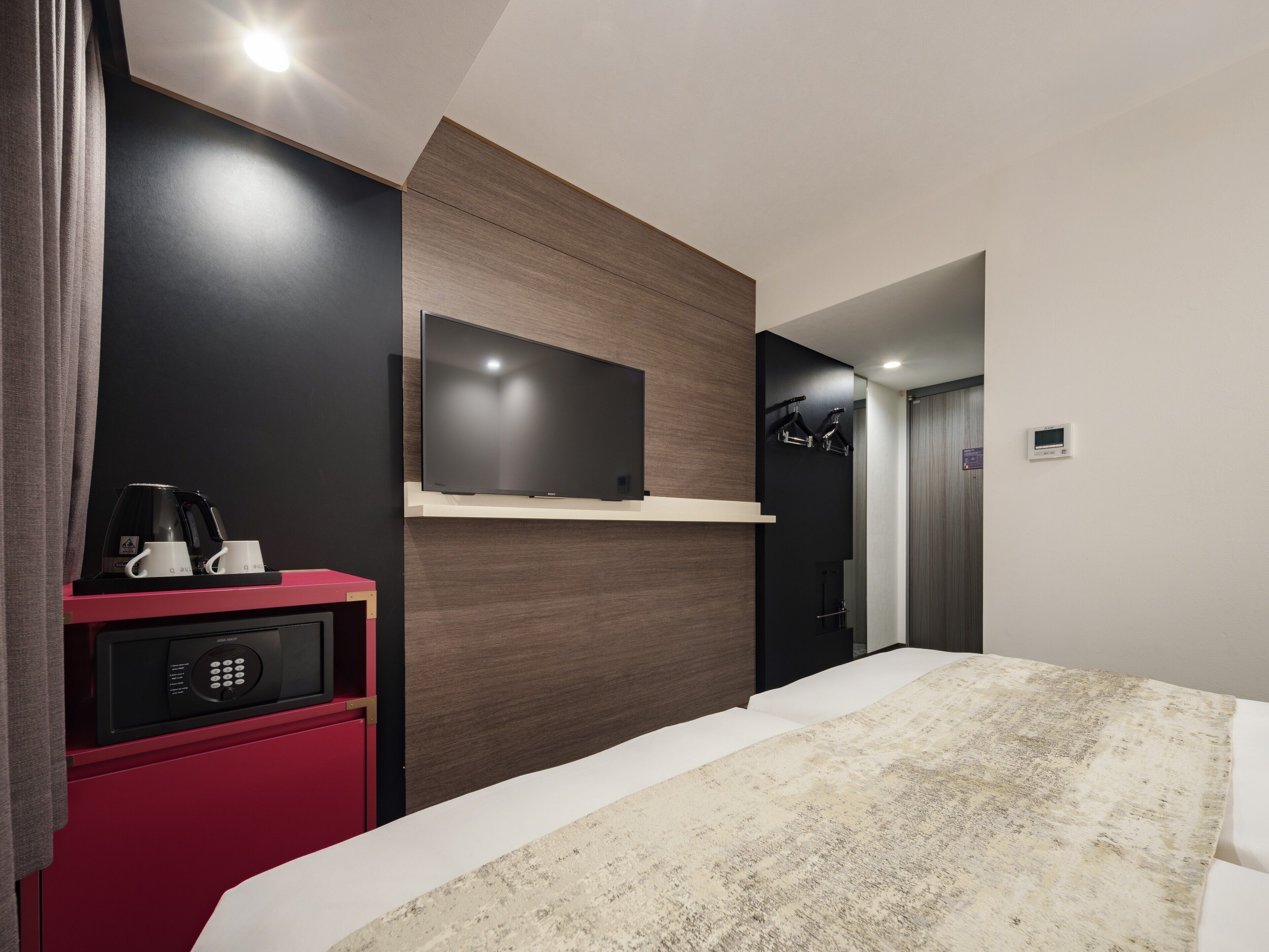 Habitación de hotel moderna con una cama grande, un televisor de pantalla plana montado en un panel de madera, un minibar con cafetera y un termostato digital en la pared.