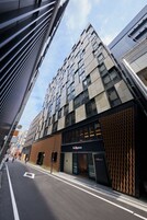A modern building with a mix of glass and brick facades, a prominent entrance with a sign, and a clear street in front.