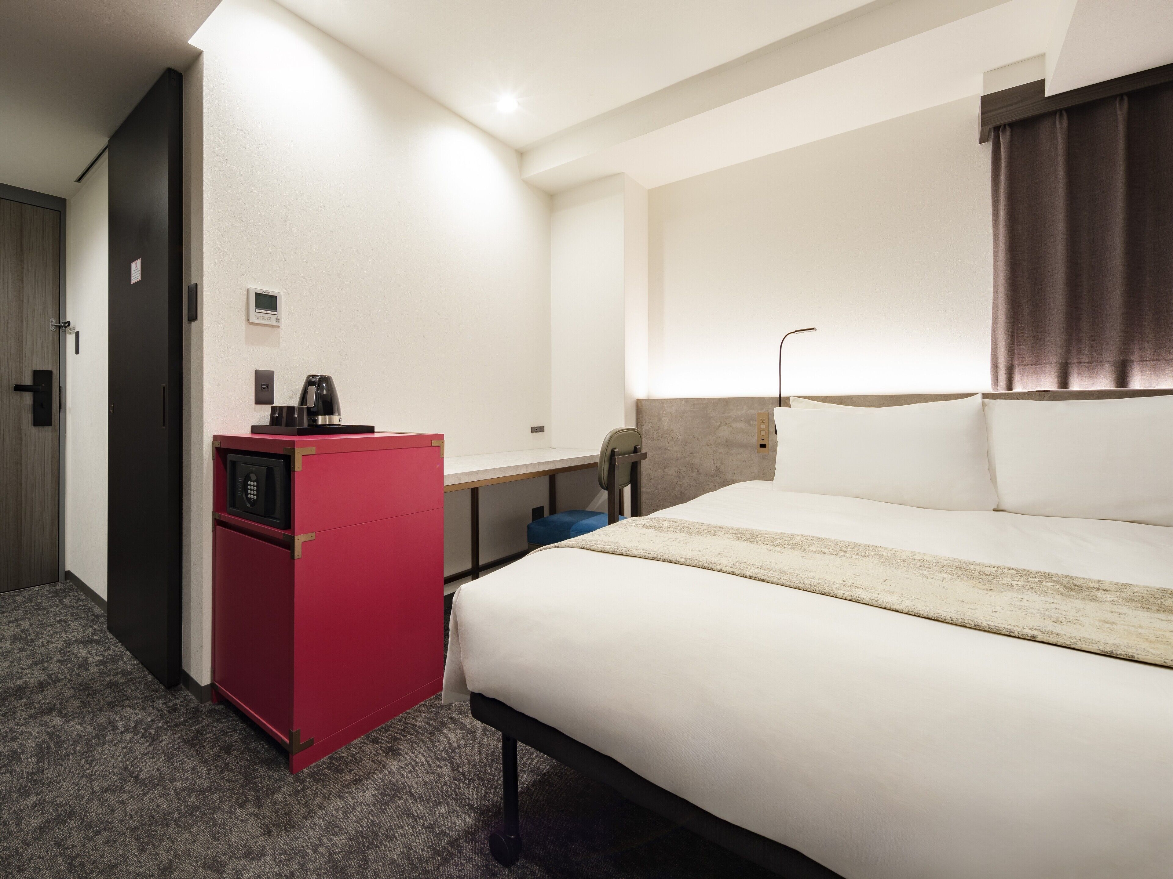 A hotel room with a bed, a desk, a red minibar, and a door.