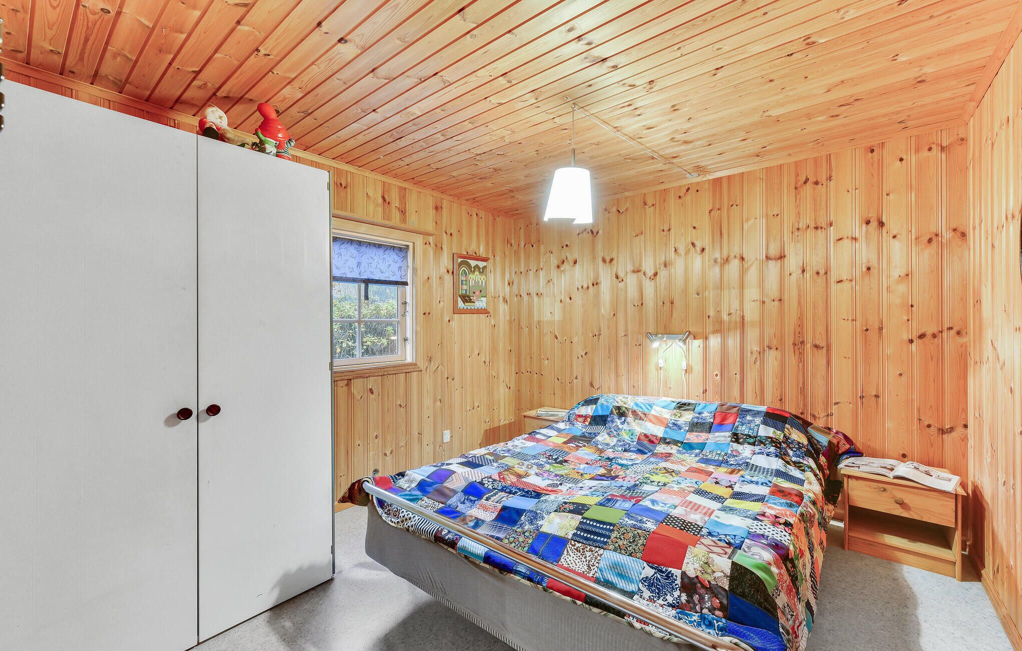 A bedroom with a wooden ceiling, a bed with a colorful quilt, a window with a view, and a white wardrobe.