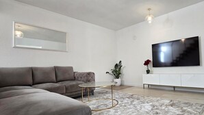 Ein modernes Wohnzimmer mit einem Ecksofa, einem runden Couchtisch, einem kleinen Beistelltisch, einer Topfpflanze und einem an der Wand befestigten Flachbildfernseher.