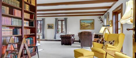 A spacious room with a high ceiling, wooden beams, a large bookshelf filled with books, a leather armchair, a yellow armchair, and a painting on the wall.
