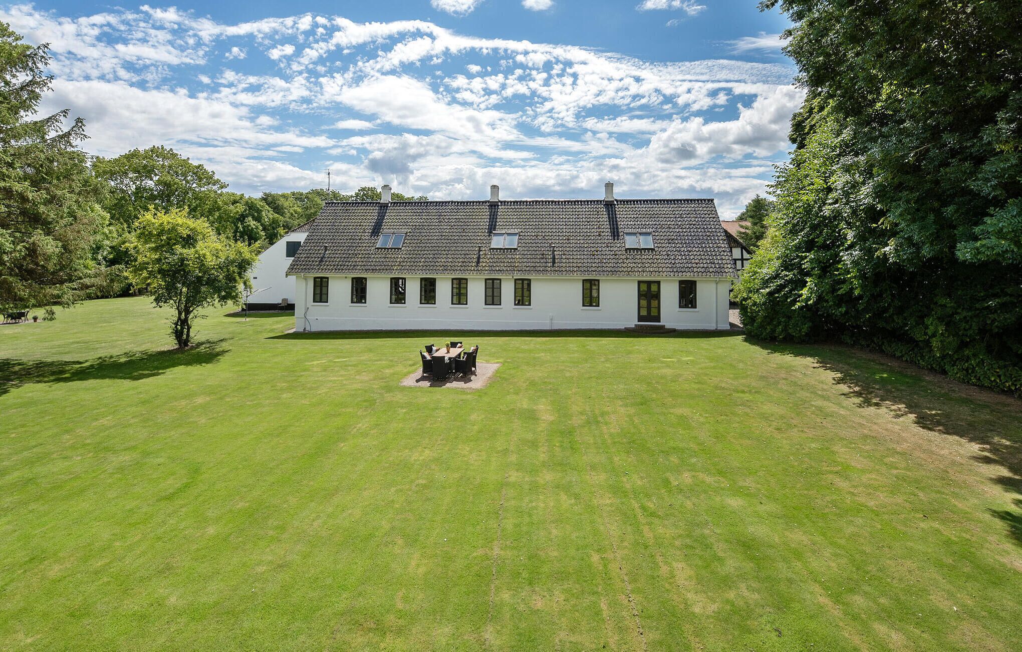 Une maison blanche à deux étages, avec un toit recouvert de tuiles, entourée d’une pelouse bien entretenue et d’arbres.