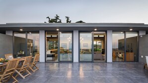 A modern house with a spacious patio, a dining area with wooden chairs, and a kitchen visible through glass doors. - Executive 9 bedroom villa with a heated pool (Daratsos)